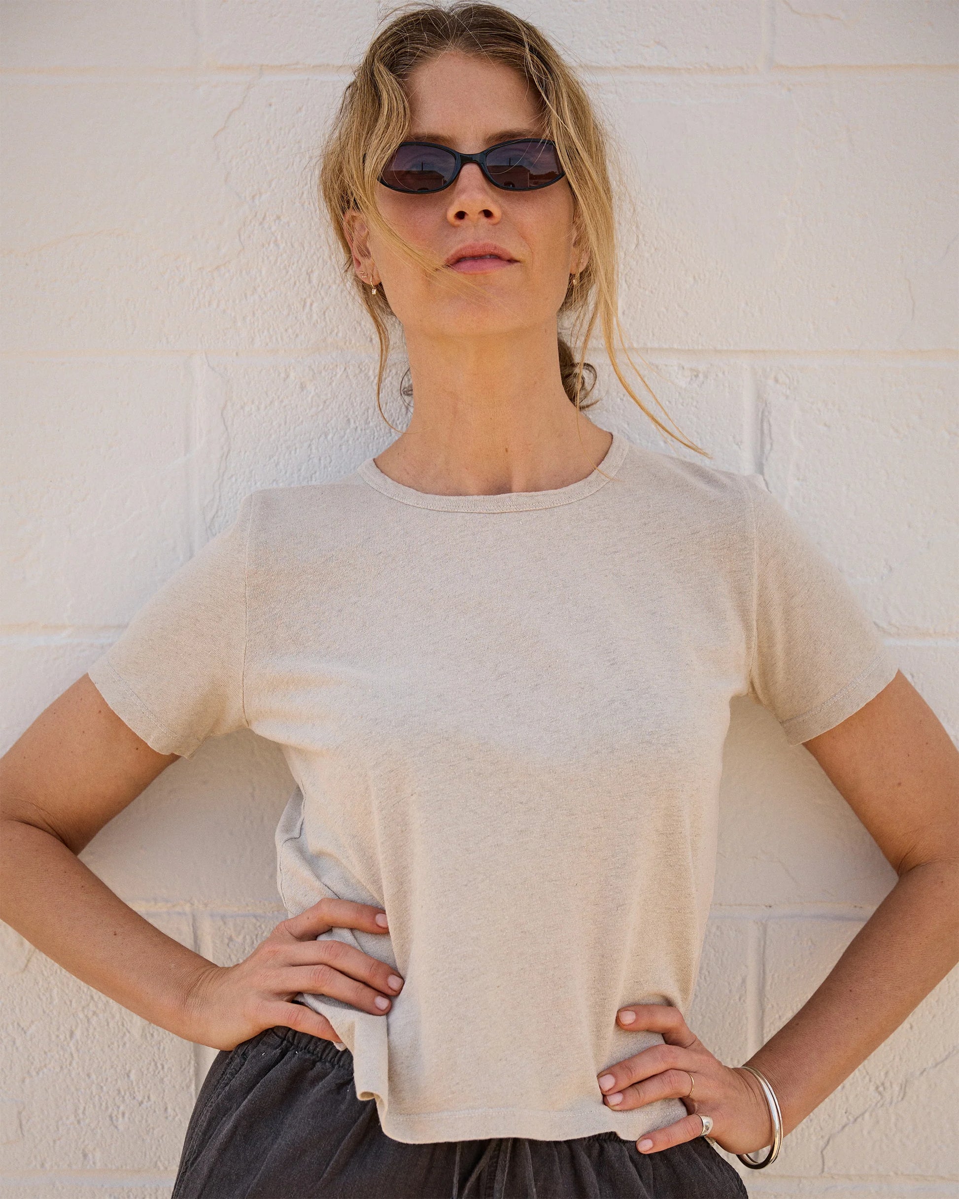 A woman in dark sunglasses, dark pants, and the LinenO Women's Sun Tee in Bone by Alex Crane stands against a white brick wall with her hands on her hips, looking confidently at the camera.