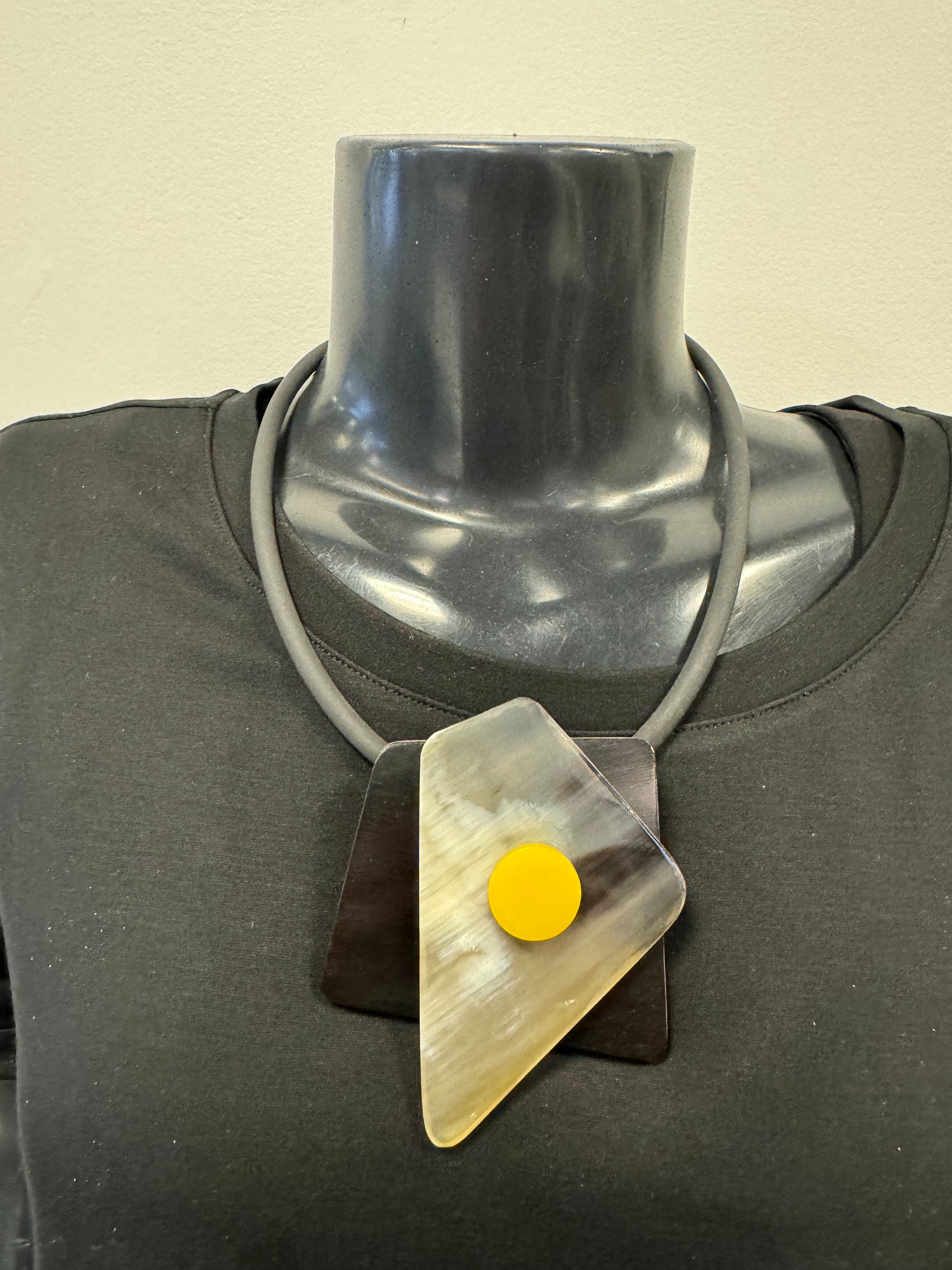 A mannequin's neck and upper chest are shown wearing a black shirt and an elegant Yellow and Black Coloured Necklace by Posies Flowers & Fashion Inc. This sophisticated accessory features abstract, layered elements in black, translucent, and yellow, creating a geometric design with triangular and circular shapes.