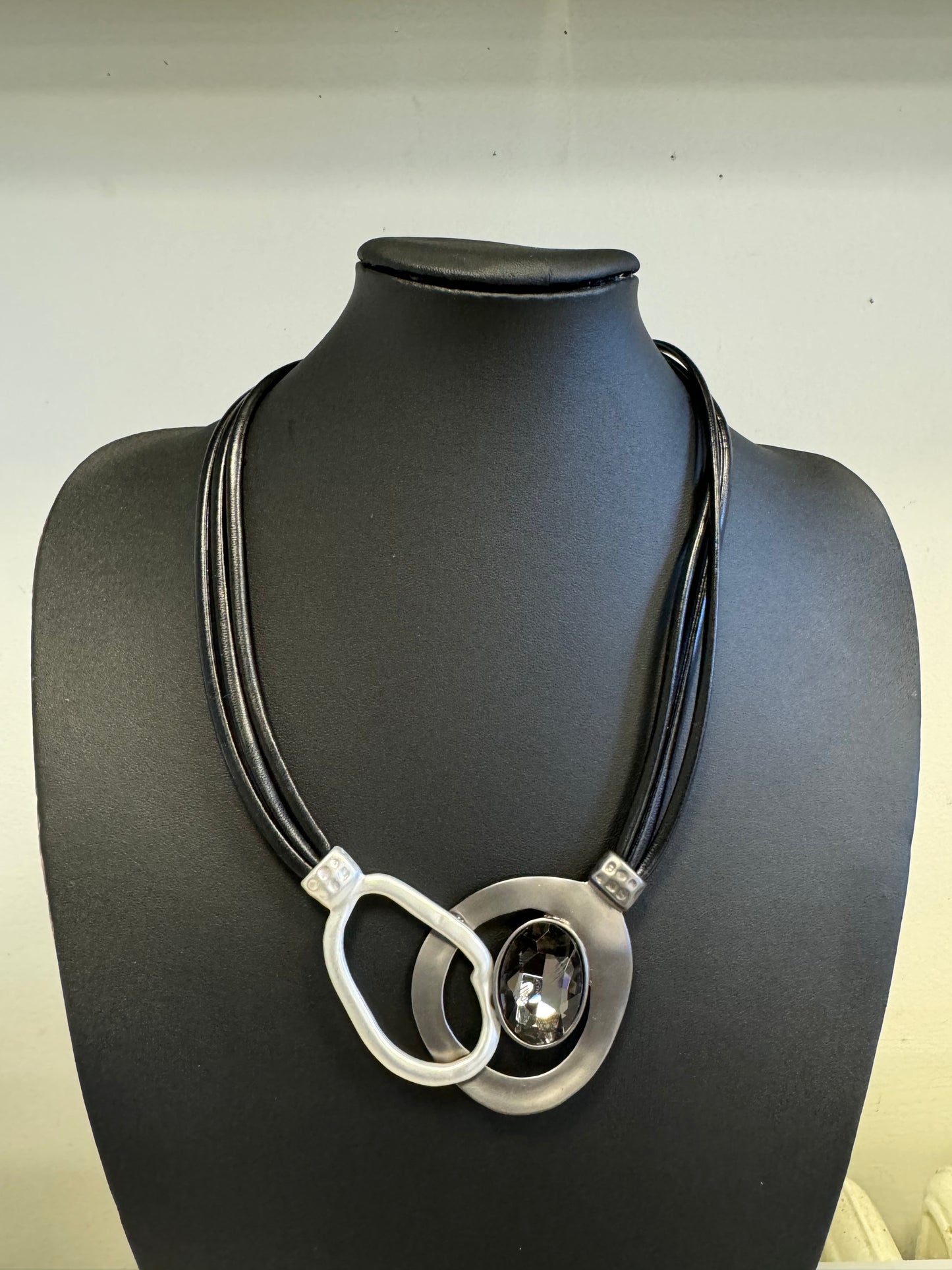 The Debbie Necklace by Elise Accessories is a captivating fashion accessory displayed on a black bust. It consists of several black cords holding two large, uniquely shaped metallic rings, with one ring featuring a striking black gemstone at its center.