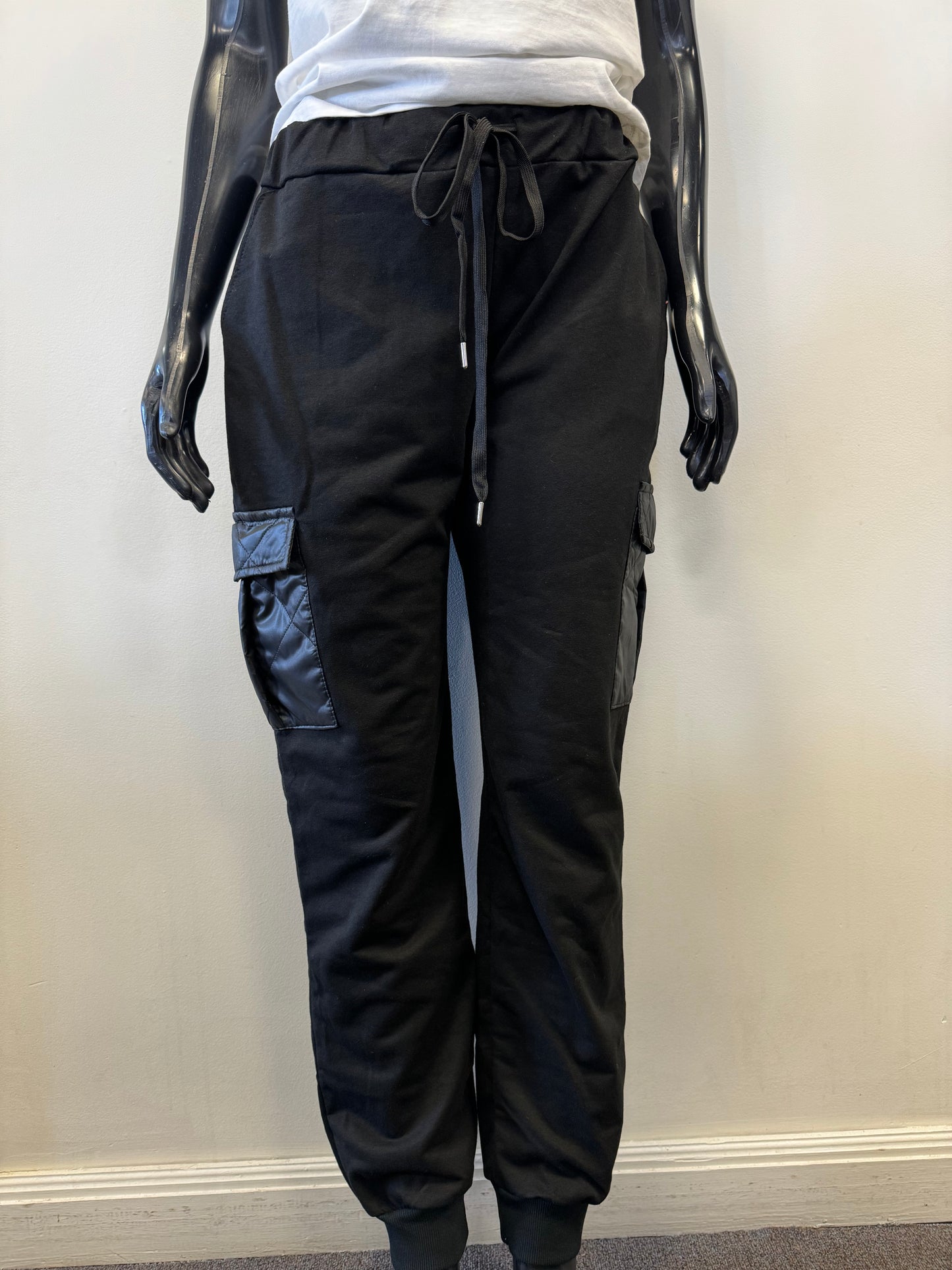 A mannequin dressed in ETERNELLE's Black Joggers with patch pockets, featuring a drawstring waist and elastic cuffs. The jogger pants, made in Italy, have stylish cargo pockets with a glossy zippered detail and are paired with a plain white top. The mannequin's hands are visible, resting by its sides.