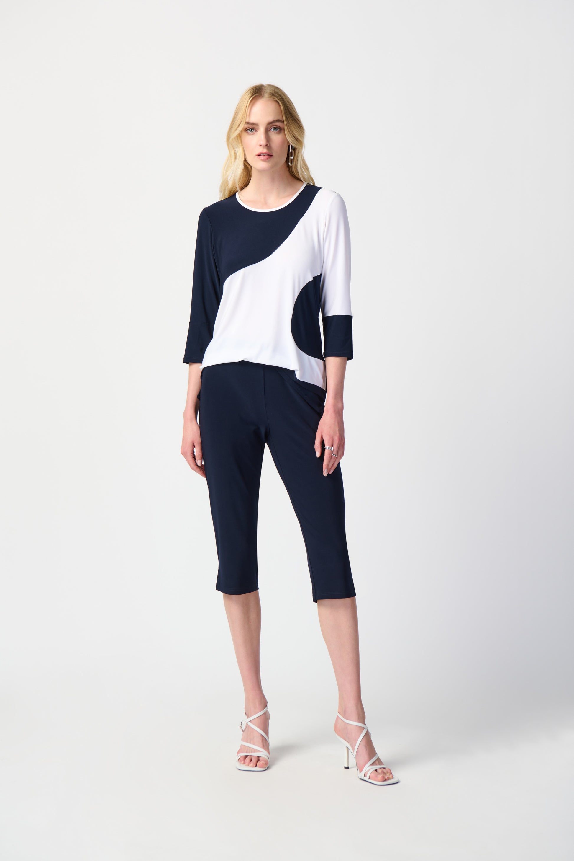 A blonde woman with loose hair wears a black and white color-blocked top featuring three-quarter sleeves, paired with Joseph Ribkoff Silky Knit Pull-On Capri Pants. She completes her look with white high-heeled sandals and silver earrings, standing against a plain white background.