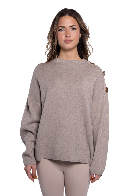 A person with long wavy brown hair is wearing a stylish top in the form of a beige, long-sleeved sweater from ETERNELLE's "Top with Golden Ornaments over the shoulder - Eternelle" collection. They stand against a white background, looking at the camera with a neutral expression. The sweater is paired effortlessly with light beige pants.