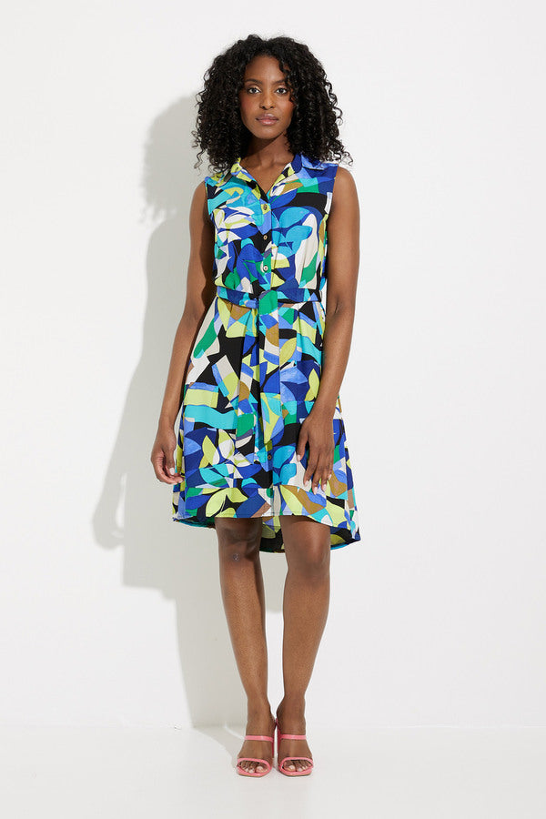 A person with curly hair stands against a white background, wearing the Joseph Ribkoff Button Front Dress Style 232090. This sleeveless dress boasts a bold geometric print perfect for any tropical getaway and is paired with pink open-toe heels. The dress features a collar and buttons down the front, as the individual stands with one hand resting by their side.