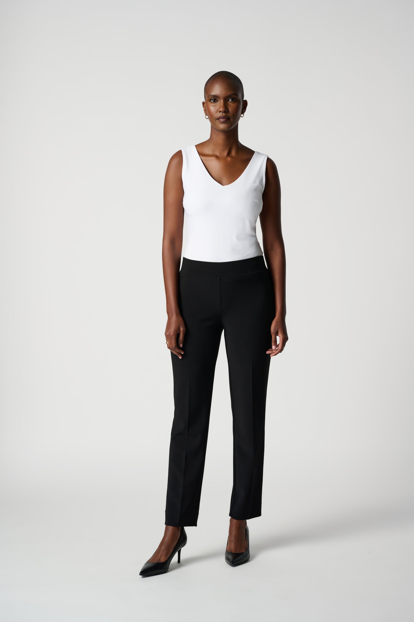 A person wearing the Classic Straight Pant in black by Joseph Ribkoff with slits at the ankles and black pointed heels, standing with their back to the camera. The person is also wearing a white top made of silky knit material. The background is plain white.