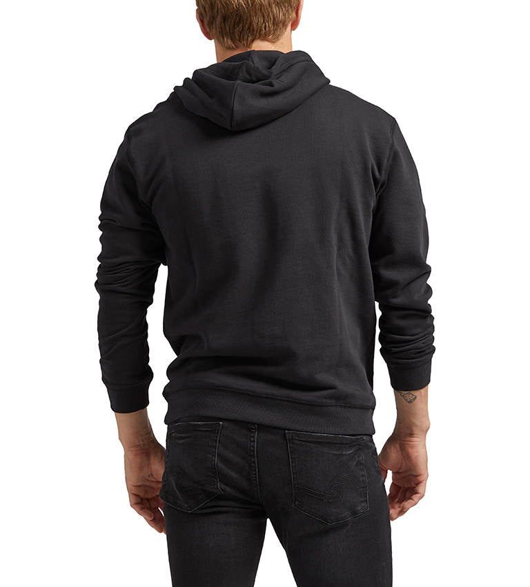 A person is wearing the Silver Jeans Co. Logo Hoodie in black, featuring "Silver Jeans Co." embroidered on the front along with geographic coordinates. The hoodie includes a front pocket and drawstrings. They are also sporting black jeans and have light facial hair.