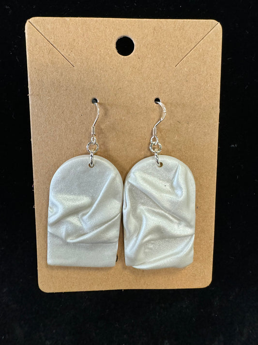 A pair of Ro&Jo Earrings 444, featuring silver, rectangular dangles with a wavy, textured surface, are displayed on a brown card with a small hole at the top for hanging. The handmade RO&JO earrings are attached to the card by silver hooks. The background is black.