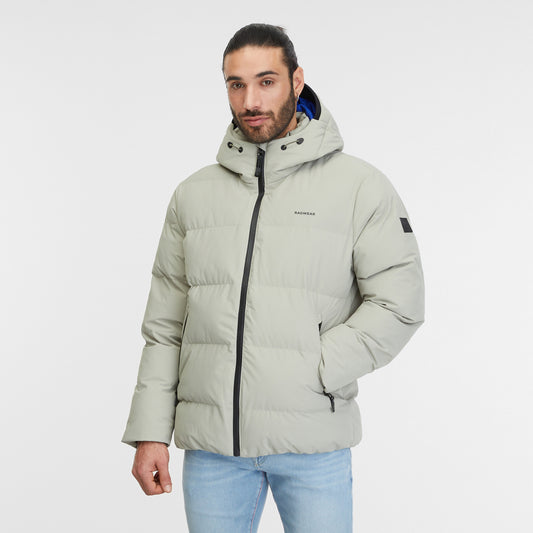 A man with long hair tied back in a bun is wearing a dusty olive Puffer Coat North from RAGWEAR and light blue jeans. He is standing against a plain white background, with his right hand in his jacket pocket and a serious expression on his face, epitomizing vegan streetwear.