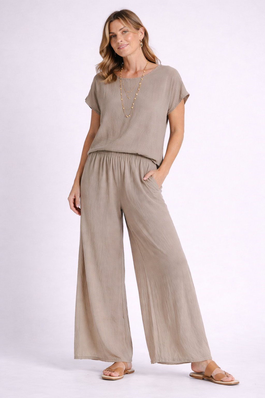 A woman smiles slightly with one hand in her pocket, wearing the loose-fitting taupe Olga Rye Top T668 and matching wide-leg pants by Oh My Gauze, styled with layered gold necklaces and tan sandals against a light background.