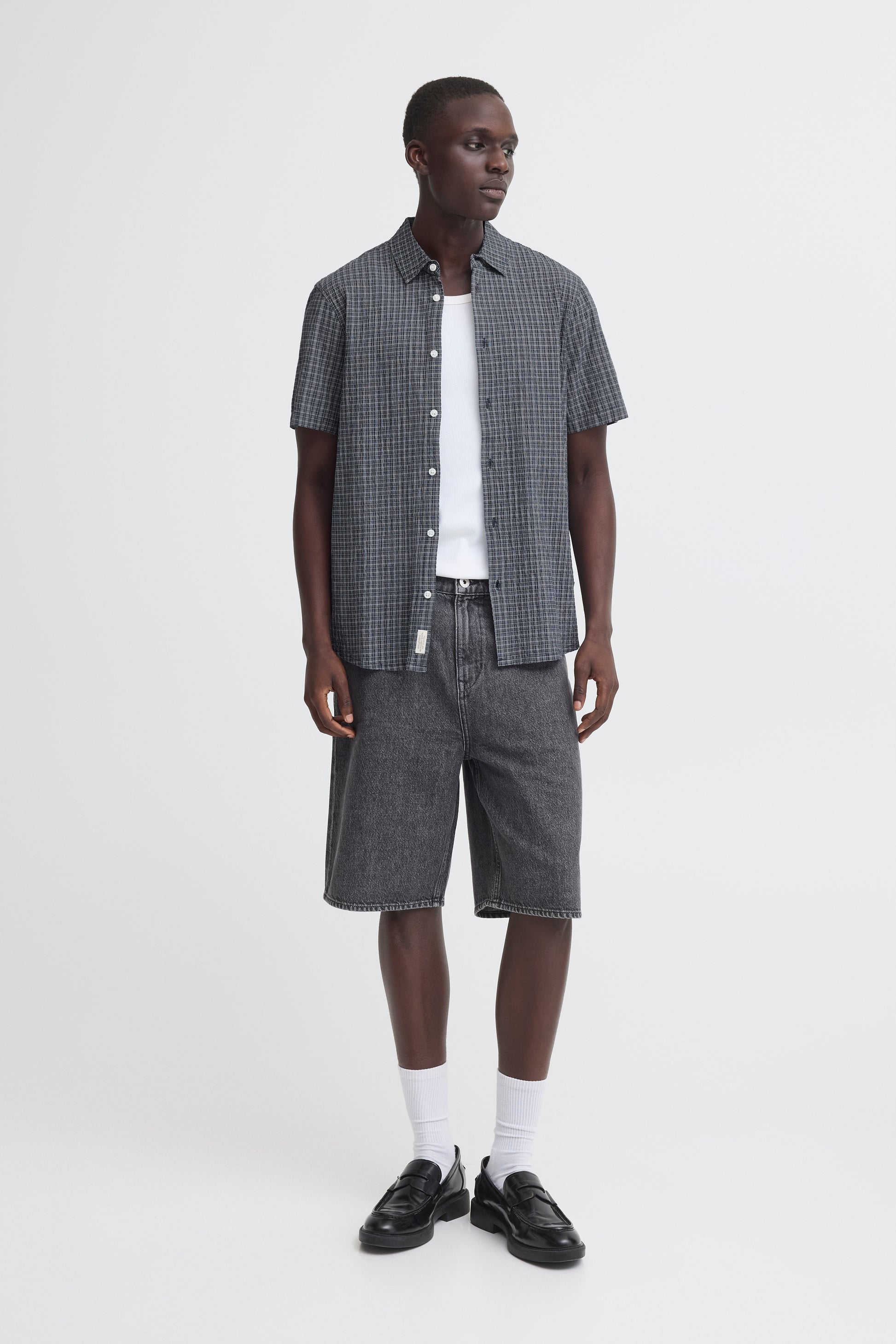 A man stands against a plain white background wearing the BLEND BARNY SHIRT 20720749-194011, a checked men's cotton shirt, over a white T-shirt with dark denim shorts, white socks, and black loafers.