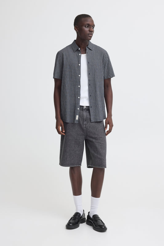 A man stands against a plain white background wearing the BLEND BARNY SHIRT 20720749-194011, a checked men's cotton shirt, over a white T-shirt with dark denim shorts, white socks, and black loafers.