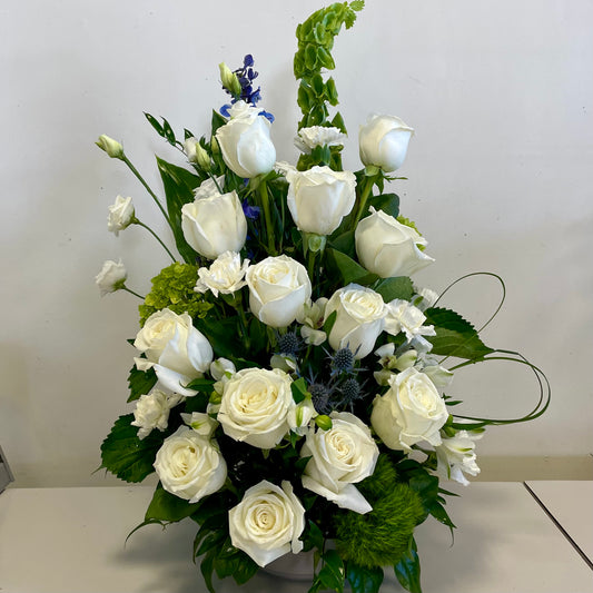 The "Pure Serenity" arrangement by Posies Flowers & Fashion offers an elegant sympathy tribute, showcasing a mix of white roses, white lilies, and green leaves beautifully presented in a white ceramic vase against a plain background.