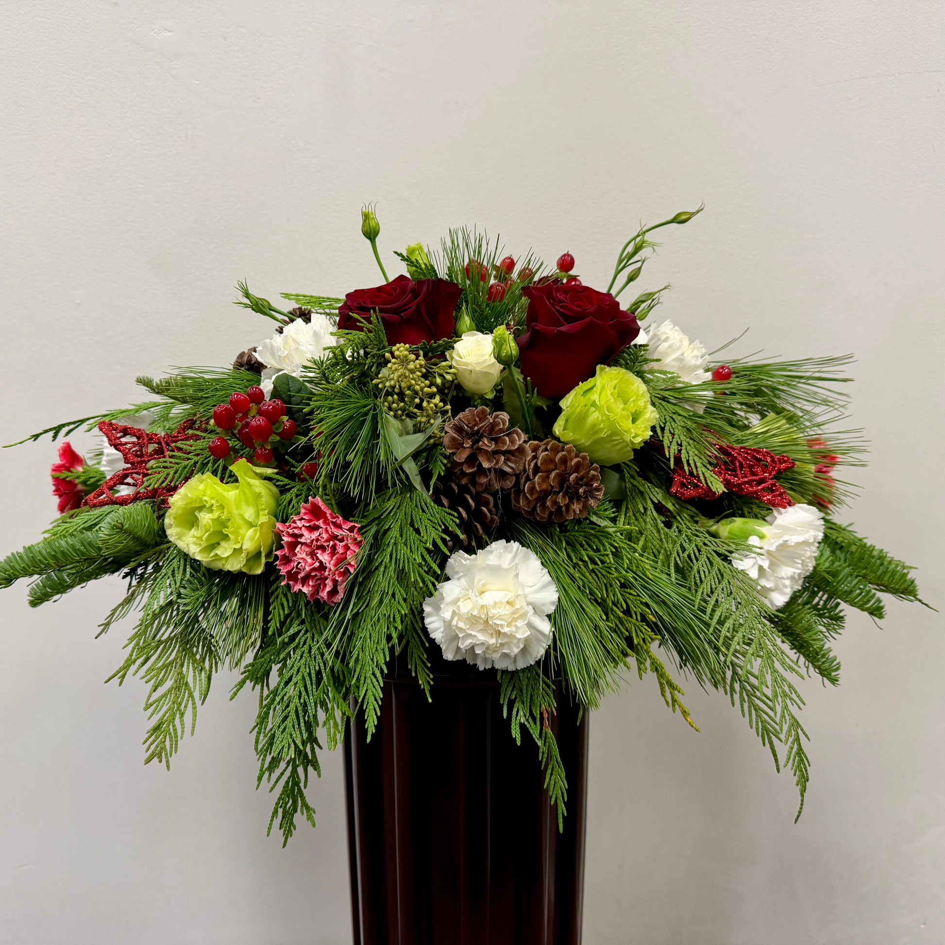 The Crimson Noel Centrepiece by Posies Flowers & Fashion features red roses, white and green carnations, red berries, pinecones, and evergreens in a dark vase—ideal as a Christmas table centrepiece against a light background.