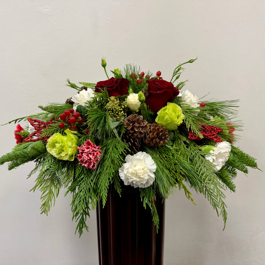 The Crimson Noel Centrepiece by Posies Flowers & Fashion features red roses, white and green carnations, red berries, pinecones, and evergreens in a dark vase—ideal as a Christmas table centrepiece against a light background.