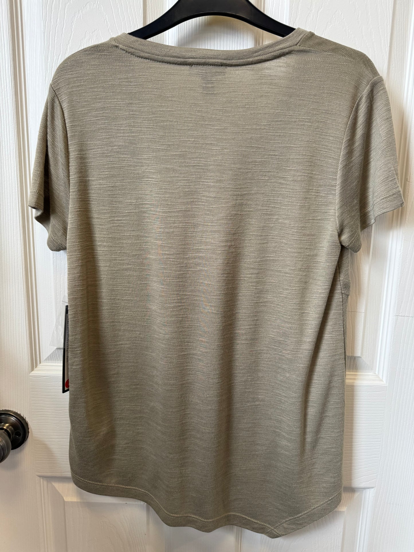 A moss green T-shirt from the brand Tribal hangs on a black hanger against a white wooden door. The shirt, part of a stylish piece, has a plain design with a soft texture. A small tag is visible on the left side, near the bottom. The doorknob on the left side of the image is black.