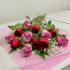 The "1 Dozen Red & Pink Roses Mixed Freshcut" bouquet by Posies Flowers & Fashion showcases a blend of red and pink roses, adorned with delicate greenery and baby's breath, elegantly wrapped in pink paper on a pristine white surface.
