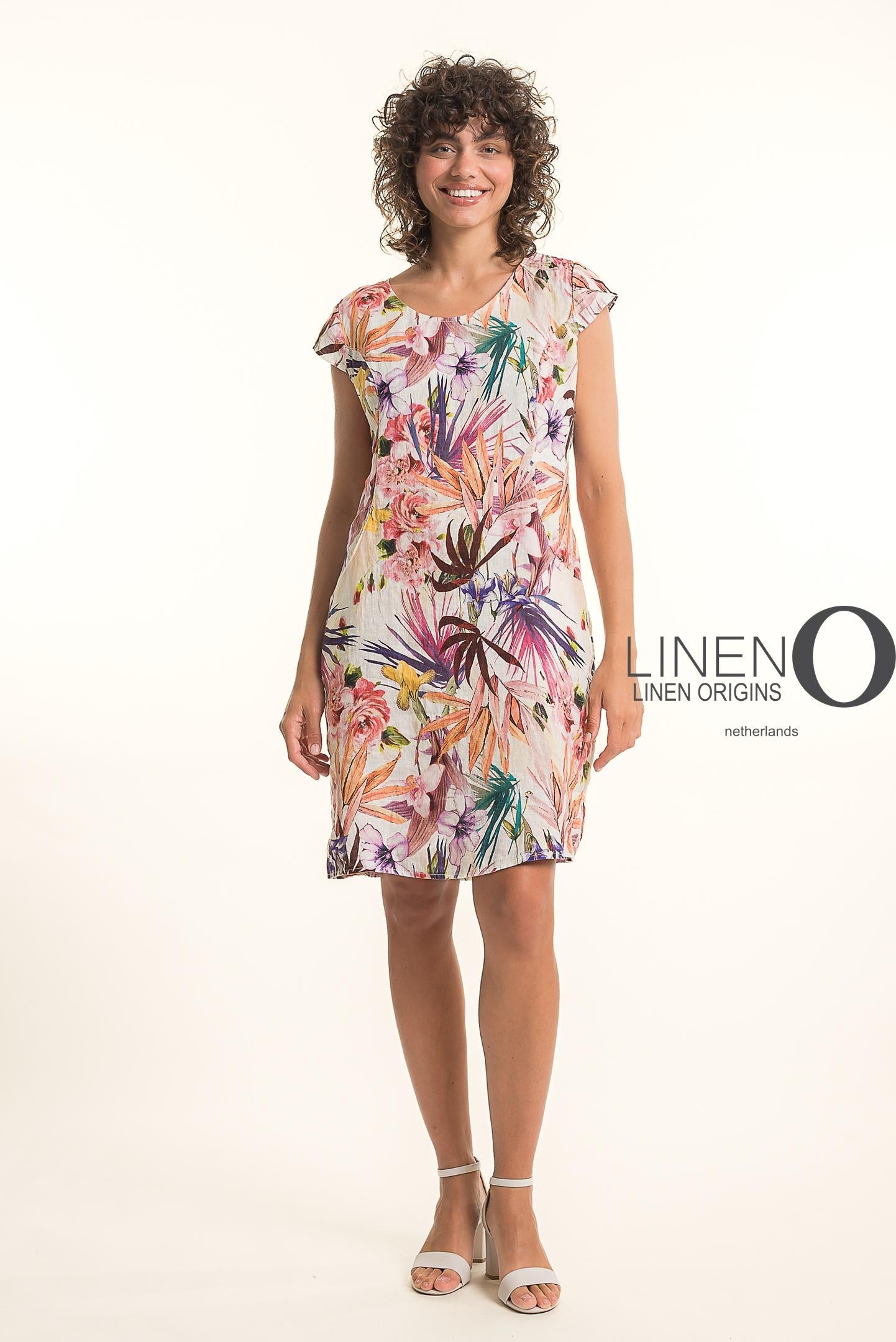 A smiling person with curly hair wears the French Linen Dress TP1758P-159 Carne by LinenO, featuring premium linen and a colorful floral and leaf print. They stand against a plain background with the "LINEN ORIGINS netherlands" logo on the right.