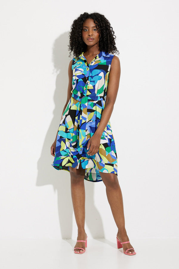 A person with curly hair stands against a white background, wearing the Joseph Ribkoff Button Front Dress Style 232090. This sleeveless dress boasts a bold geometric print perfect for any tropical getaway and is paired with pink open-toe heels. The dress features a collar and buttons down the front, as the individual stands with one hand resting by their side.