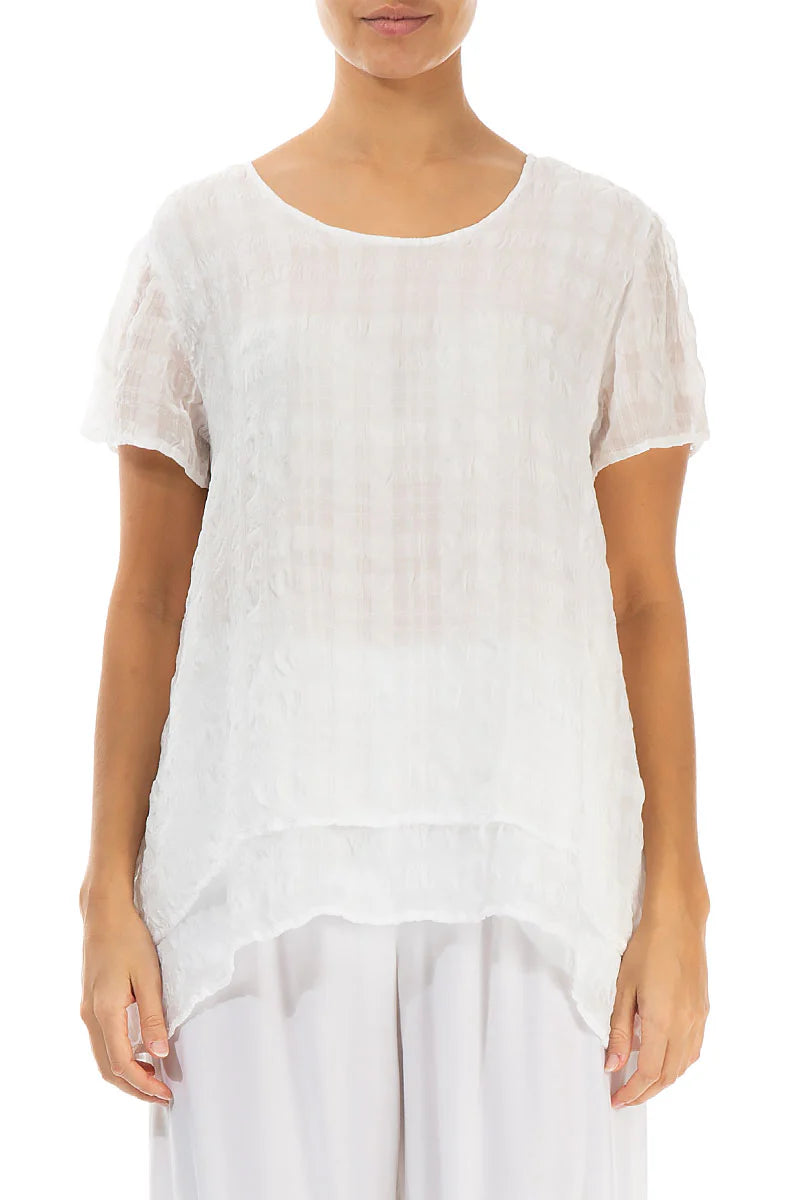 A person wearing the Paolo Tricot LAYERED WHITE LIGHT SILK BLOUSE by GRIZAS, featuring a loose fit, short sleeves, round neckline, and asymmetrical hem, paired with white pants against a plain white background.