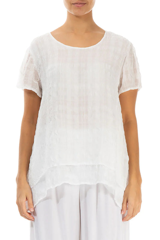A person wearing the Paolo Tricot LAYERED WHITE LIGHT SILK BLOUSE by GRIZAS, featuring a loose fit, short sleeves, round neckline, and asymmetrical hem, paired with white pants against a plain white background.