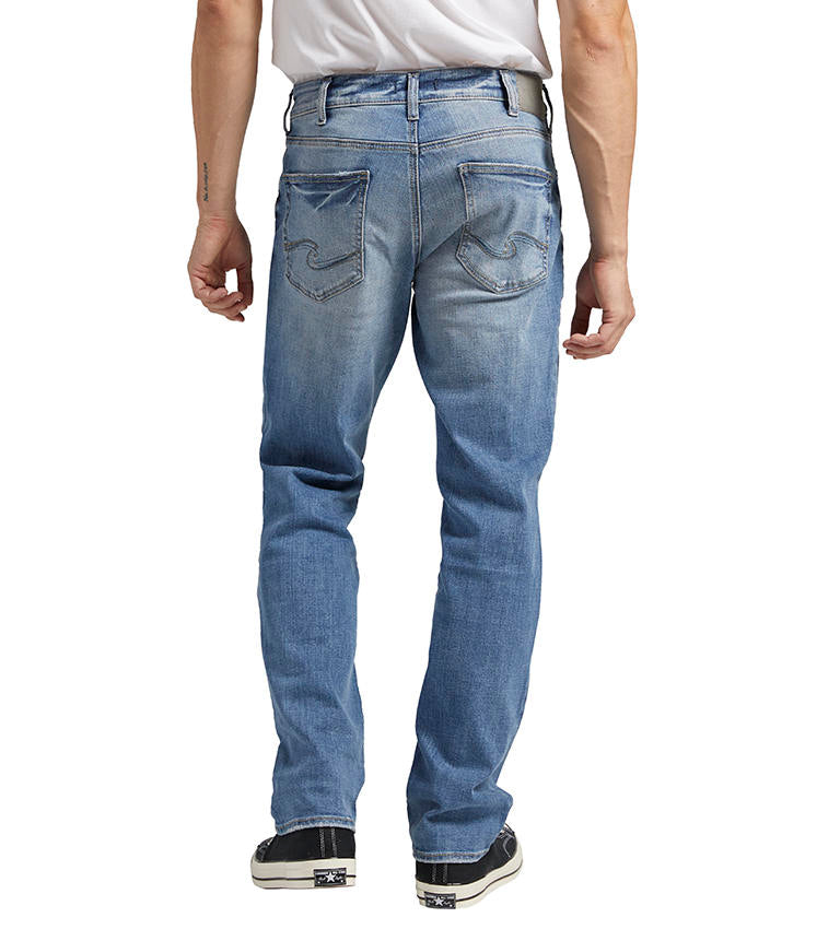 A person is wearing a white shirt tucked into a pair of Grayson Classic Fit Straight Leg Jeans by Silver Jeans Co. These indigo denim men's jeans feature a straight-leg cut with a faded design on the thighs and knees. They are also wearing black and white low-top sneakers.