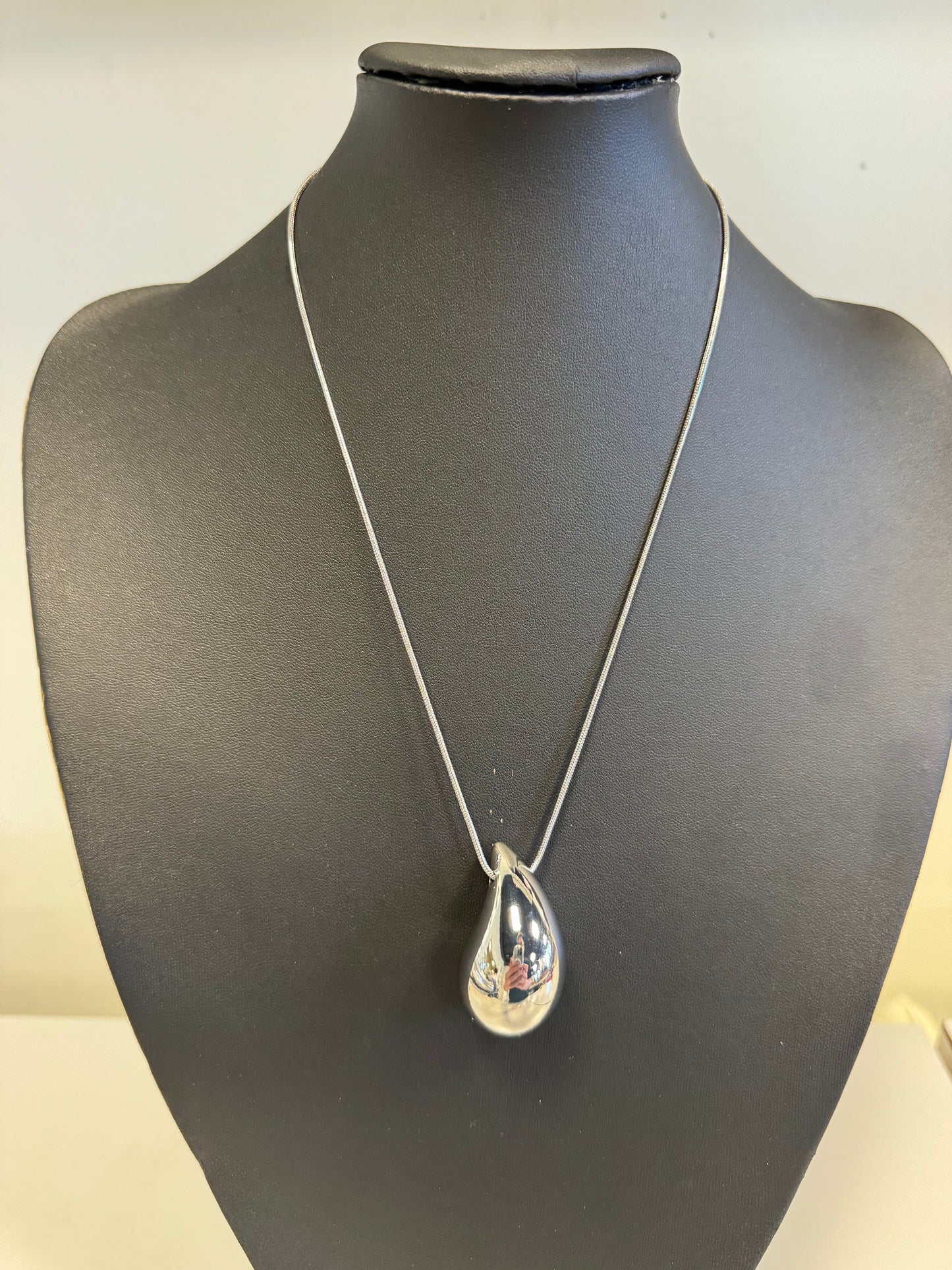 The Andie 60 Necklace by Elise Accessories features a sleek silver teardrop-shaped pendant elegantly adorning a thin chain, beautifully showcased on a black bust. Its shiny, reflective surface serves as a captivating centerpiece for any collection from Elise Accessories.