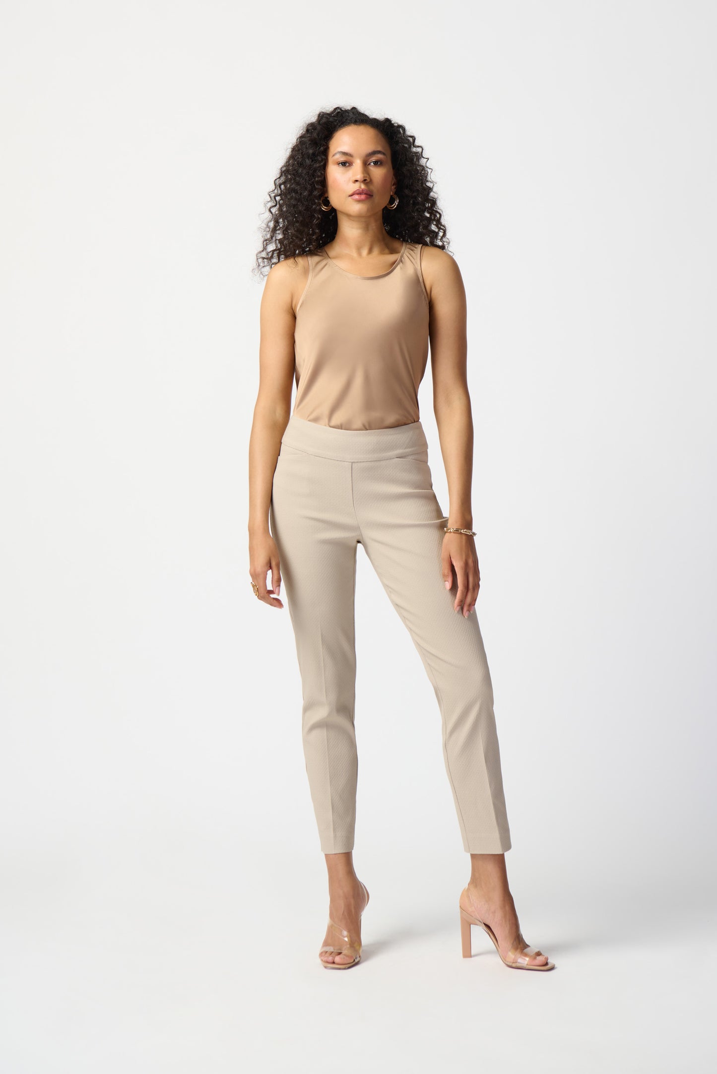 A woman with curly hair is standing against a plain white background. She is wearing a sleeveless beige top, Joseph Ribkoff Jacquard Pull-On Pants with a structured contour waistband in light beige, and clear high-heeled shoes. She is looking confidently at the camera, with one hand resting by her side and the other hanging loosely.