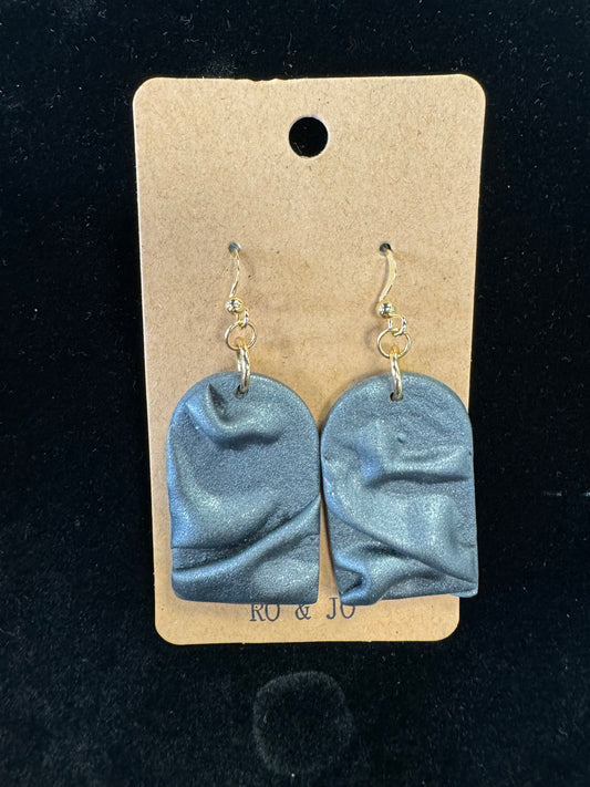 A pair of Ro&Jo Earrings 440, handmade from polymer clay with a textured, shiny gray finish, is displayed on a brown rectangular card with a small hole at the top. The RO&JO earrings feature uneven surfaces that create a wavy appearance and hang from gold hooks. The background is black.