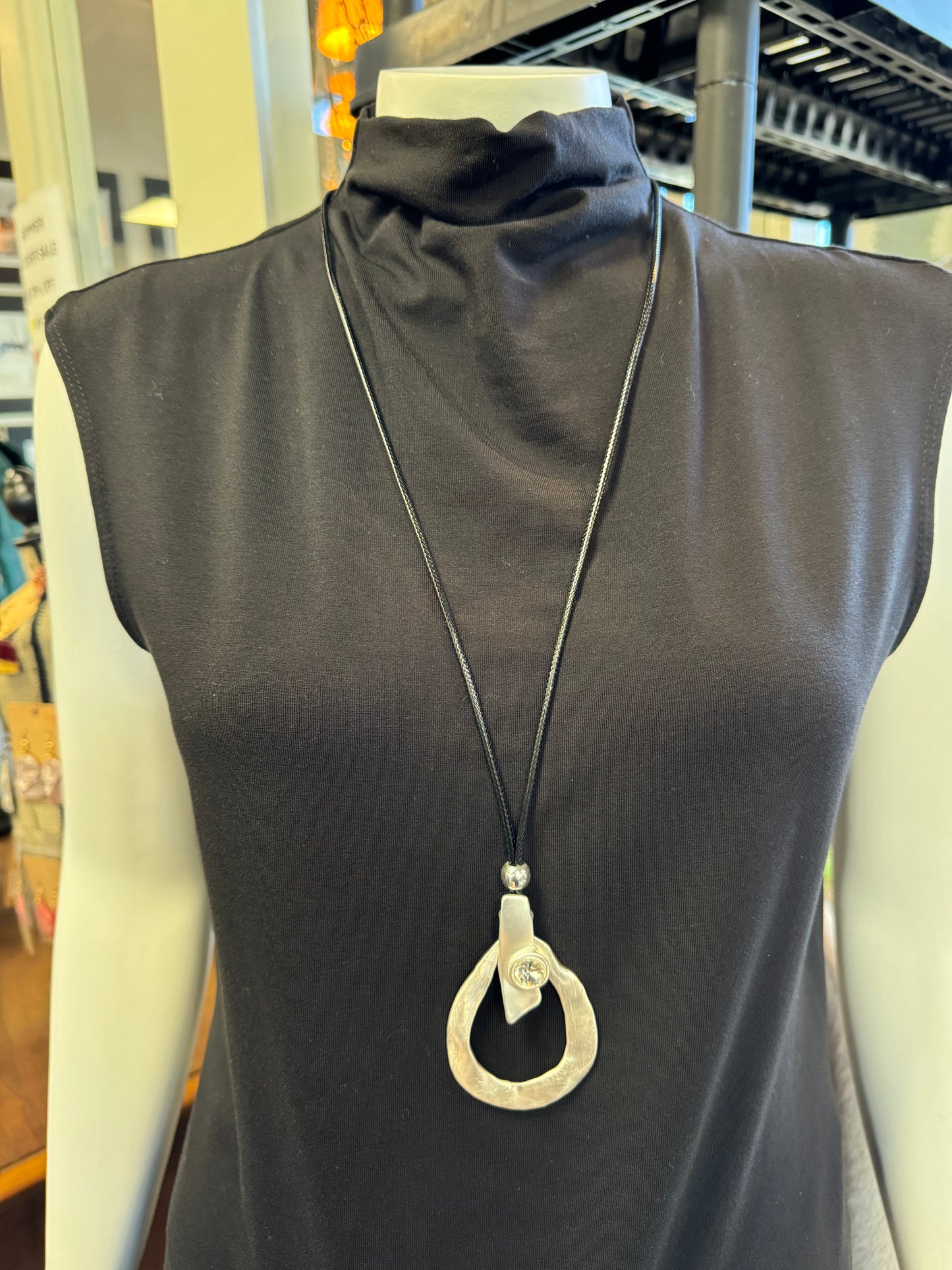 In an elegant display, a mannequin dons a sleeveless black turtleneck top featuring the striking piece, Necklace 3 - Elise Accessories from Elise Accessories: a long black necklace adorned with a large, silver teardrop-shaped pendant. The backdrop includes store fixtures and displays from the jewelry collection.