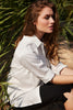 A woman with long hair sits outdoors among greenery, wearing SUNDAY's fashion-forward Top 6331 and black pants. She appears relaxed, savoring a calm Sunday, with her eyes closed and sunlight illuminating her face.