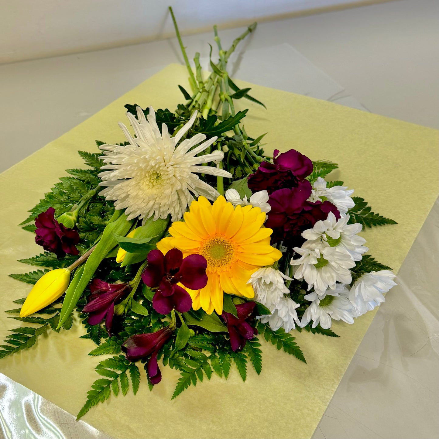 The "Sweet Harmony" bouquet by Posies Flowers & Fashion features a vibrant mix of yellow and white chrysanthemums, dark red daisies, and lush green foliage beautifully laid out on a pale yellow sheet. The arrangement is exquisitely designed against a neutral background. Please note that substitutions may occur based on seasonal availability.