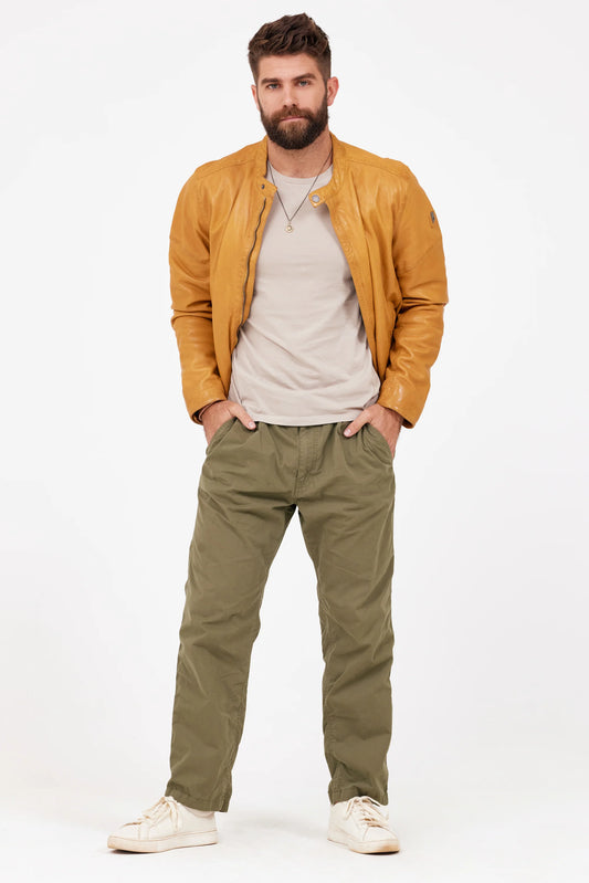 A bearded man is standing against a white background, wearing the MAURITIUS LEATHER Solvic CF Leather Jacket in Curry over a light gray T-shirt, accompanied by olive green cargo pants and white sneakers. His hands are in his pockets and he has a neutral expression on his face. The lightweight outfit contrasts sharply with the navy backdrop behind him.