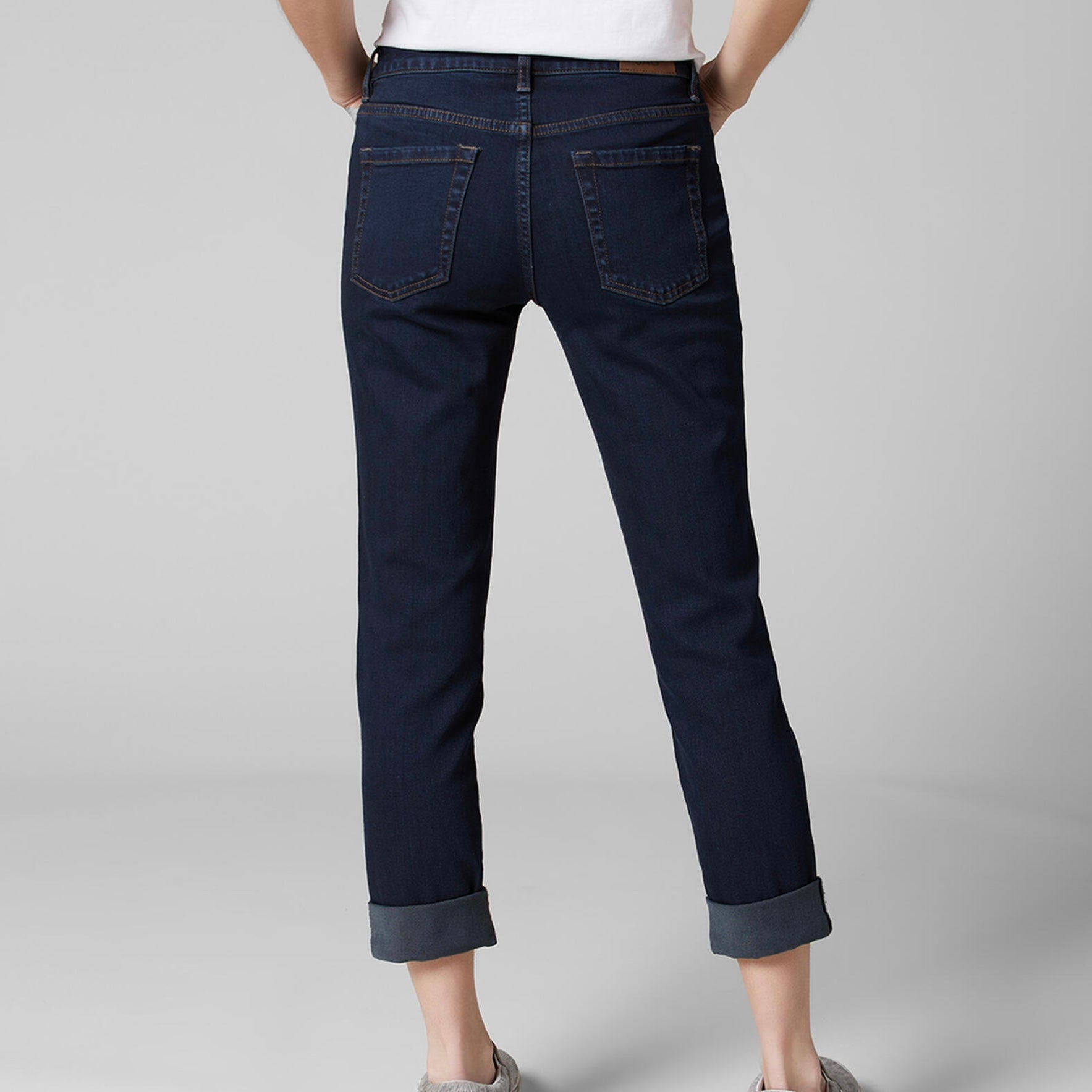 A person stands against a plain grey background wearing a white V-neck T-shirt, dark blue Carter Mid Rise Girlfriend Jeans by JAG made from Sustainable Premium Stretch denim with rolled cuffs, and has hands resting at sides. The person has long, straight hair.