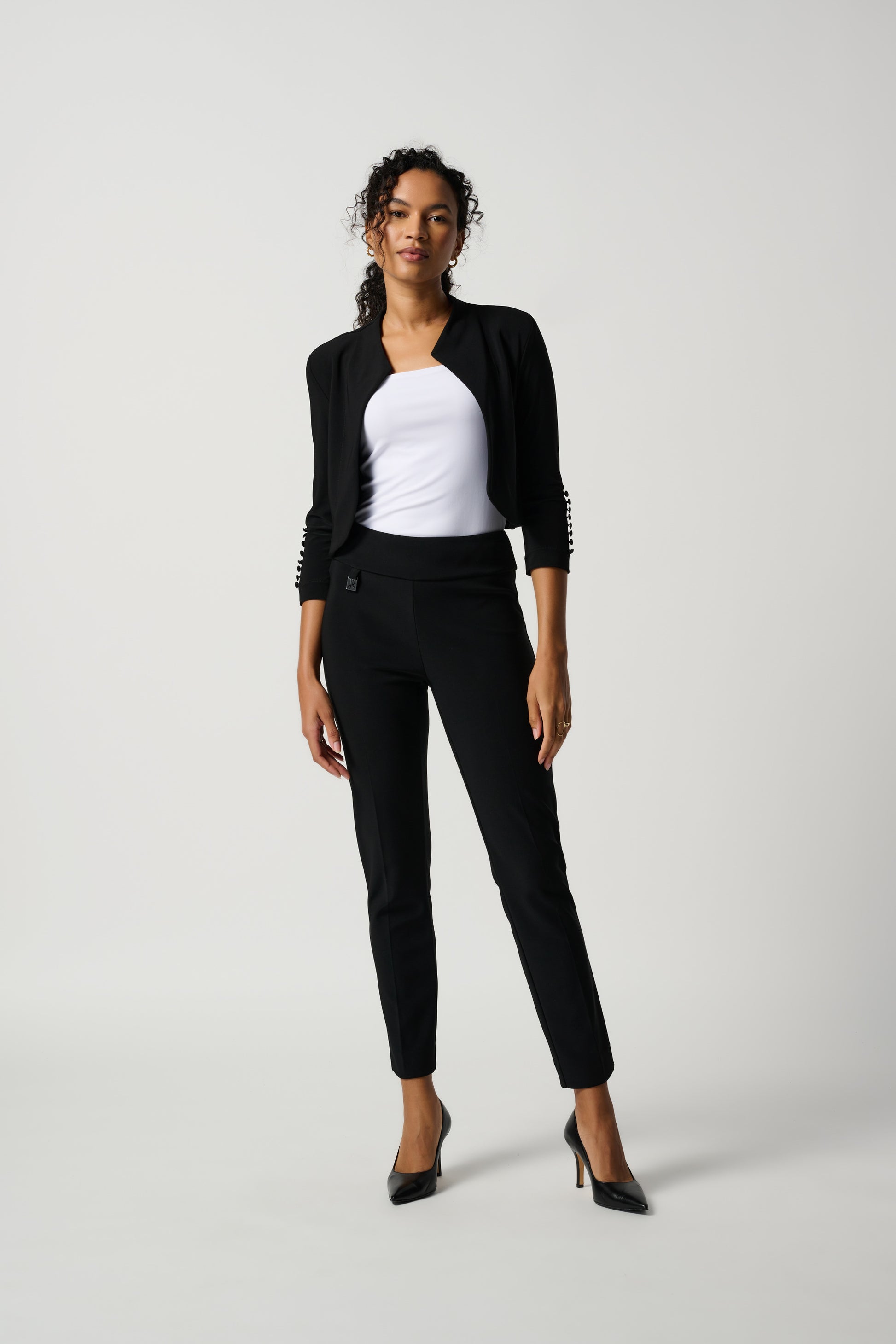 A person is standing and posing in Joseph Ribkoff's Classic Tailored Slim Pant (144092NOS-6), made from flattering silky knit fabric, paired with black high-heeled shoes. They are wearing a white top. The background is a plain light color.