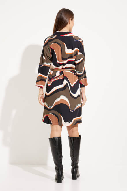 A woman stands against a white background, wearing the Abstract Print Shirt Dress Style 233224 by Joseph Ribkoff, which features wavy, multicolored patterns in beige, brown, and white. The long-sleeve dress has a belted waist and buttons down the top half. She is also wearing black knee-high boots.