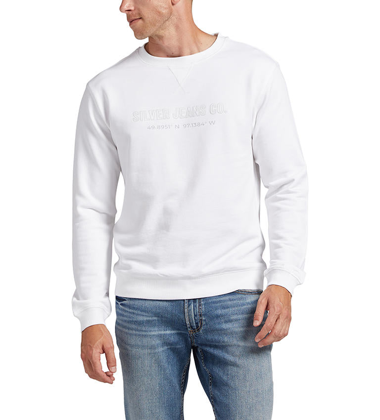 A man is modeling the Silver Jeans Co. Mens Crewneck Sweatshirt in white, featuring subtle embroidered text "49.8951°N 97.1384°W" on the front. He pairs it with light blue jeans, has short hair, and is looking to his left against a plain white background.