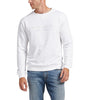 A man is modeling the Silver Jeans Co. Mens Crewneck Sweatshirt in white, featuring subtle embroidered text "49.8951°N 97.1384°W" on the front. He pairs it with light blue jeans, has short hair, and is looking to his left against a plain white background.