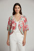 A woman wears the TaraVao Top Megan 5098 with pants and a loose, colorful floral kimono-style jacket in red, green, and pink. She smiles softly against a neutral background. Made in Bali by TaraVao.