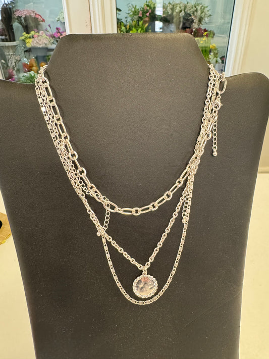 The Simple Silver Coloured Necklace from Posies Flowers & Fashion Inc. is displayed on a black stand, showcasing three different chains of varying lengths, with the shortest chain adorned by a round pendant. This elegant piece shines against a backdrop of blurred floral arrangements, epitomizing timeless jewelry.