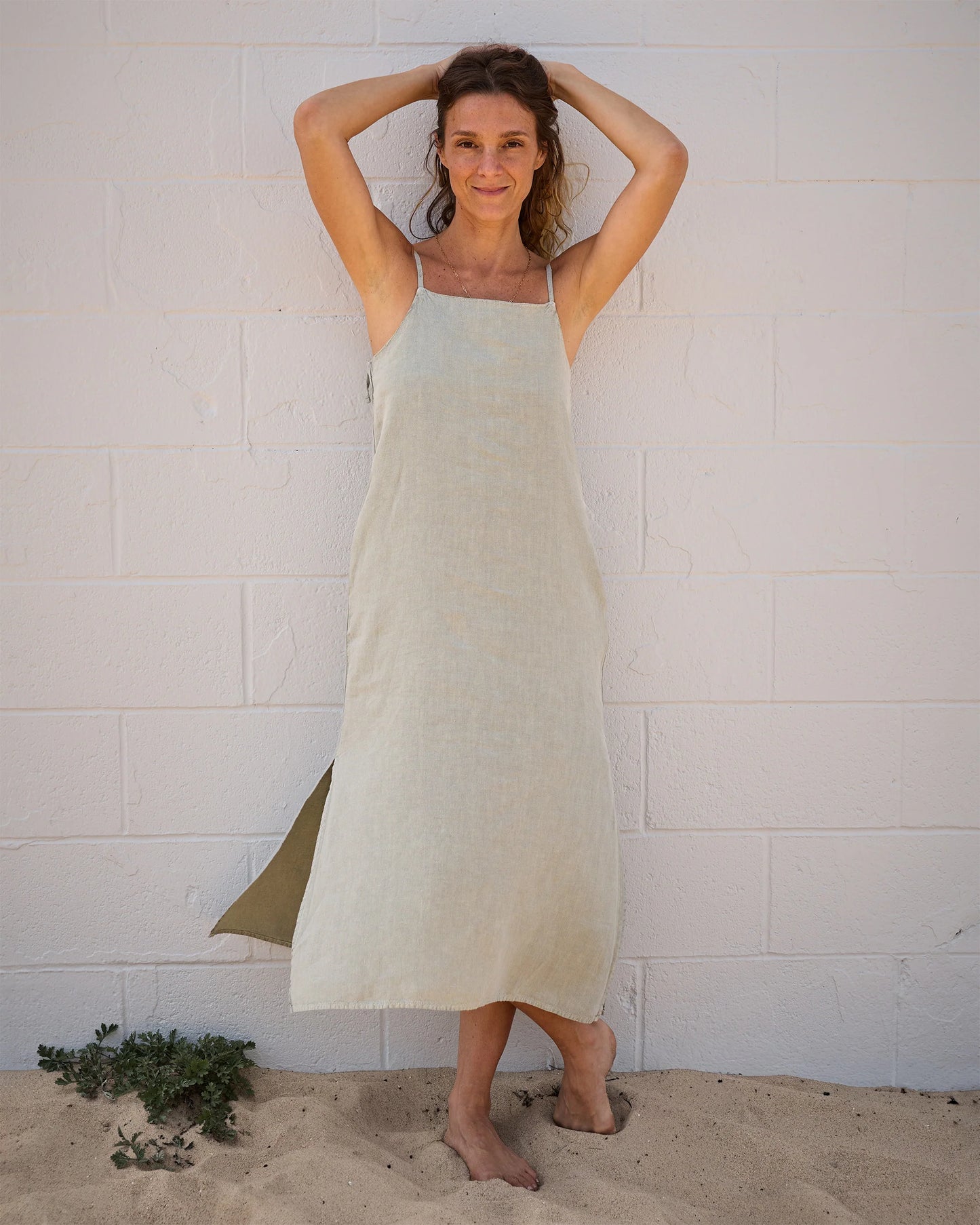 A woman stands barefoot on sand by a white brick wall, smiling with her arms raised. She wears the Women's Penny Slip in Seagrass linen by Alex Crane. Small green plants are visible in the sand near her feet.