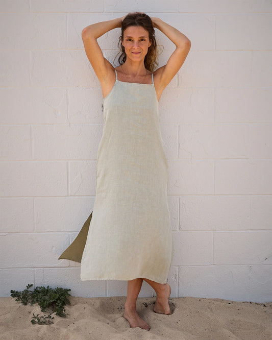 A woman stands barefoot on sand by a white brick wall, smiling with her arms raised. She wears the Women's Penny Slip in Seagrass linen by Alex Crane. Small green plants are visible in the sand near her feet.
