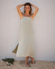 A woman stands barefoot on sand by a white brick wall, smiling with her arms raised. She wears the Women's Penny Slip in Seagrass linen by Alex Crane. Small green plants are visible in the sand near her feet.