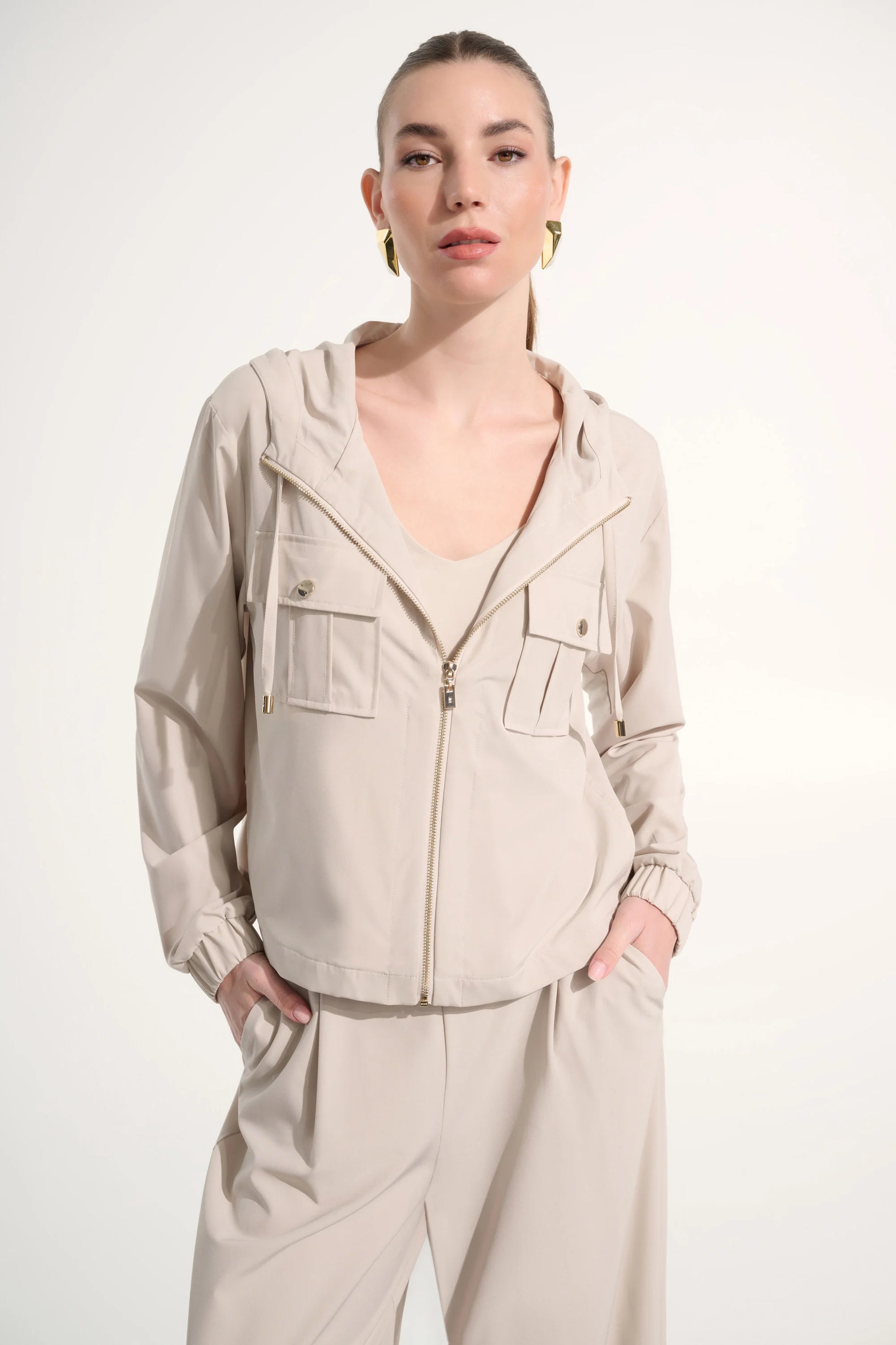 A woman with slicked-back hair models the Joseph Ribkoff Stretch Woven Hooded Boxy Utility Jacket 262621 in light beige, featuring chest pockets and paired with wide-leg pants. She accessorizes with gold hoops, perfect for casual wear.