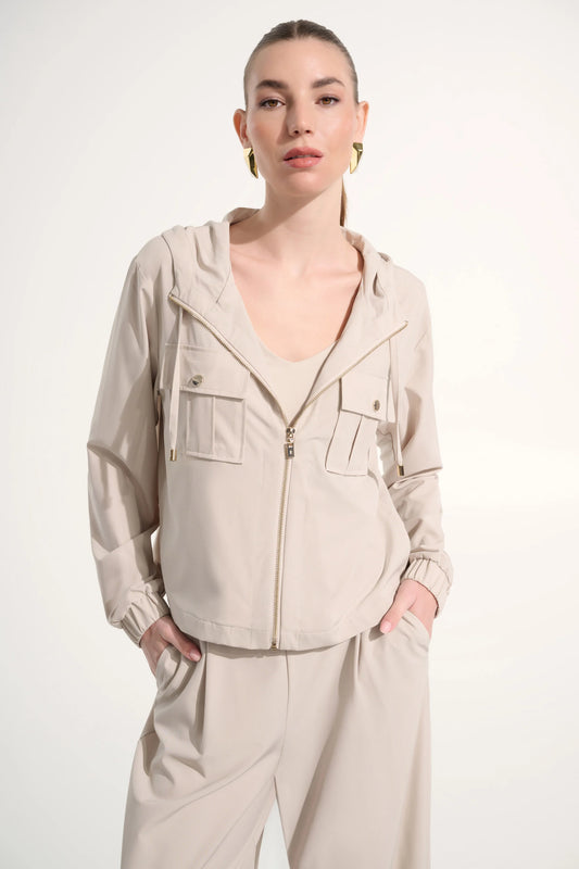 A woman with slicked-back hair models the Joseph Ribkoff Stretch Woven Hooded Boxy Utility Jacket 262621 in light beige, featuring chest pockets and paired with wide-leg pants. She accessorizes with gold hoops, perfect for casual wear.