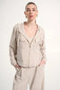 A woman with slicked-back hair models the Joseph Ribkoff Stretch Woven Hooded Boxy Utility Jacket 262621 in light beige, featuring chest pockets and paired with wide-leg pants. She accessorizes with gold hoops, perfect for casual wear.