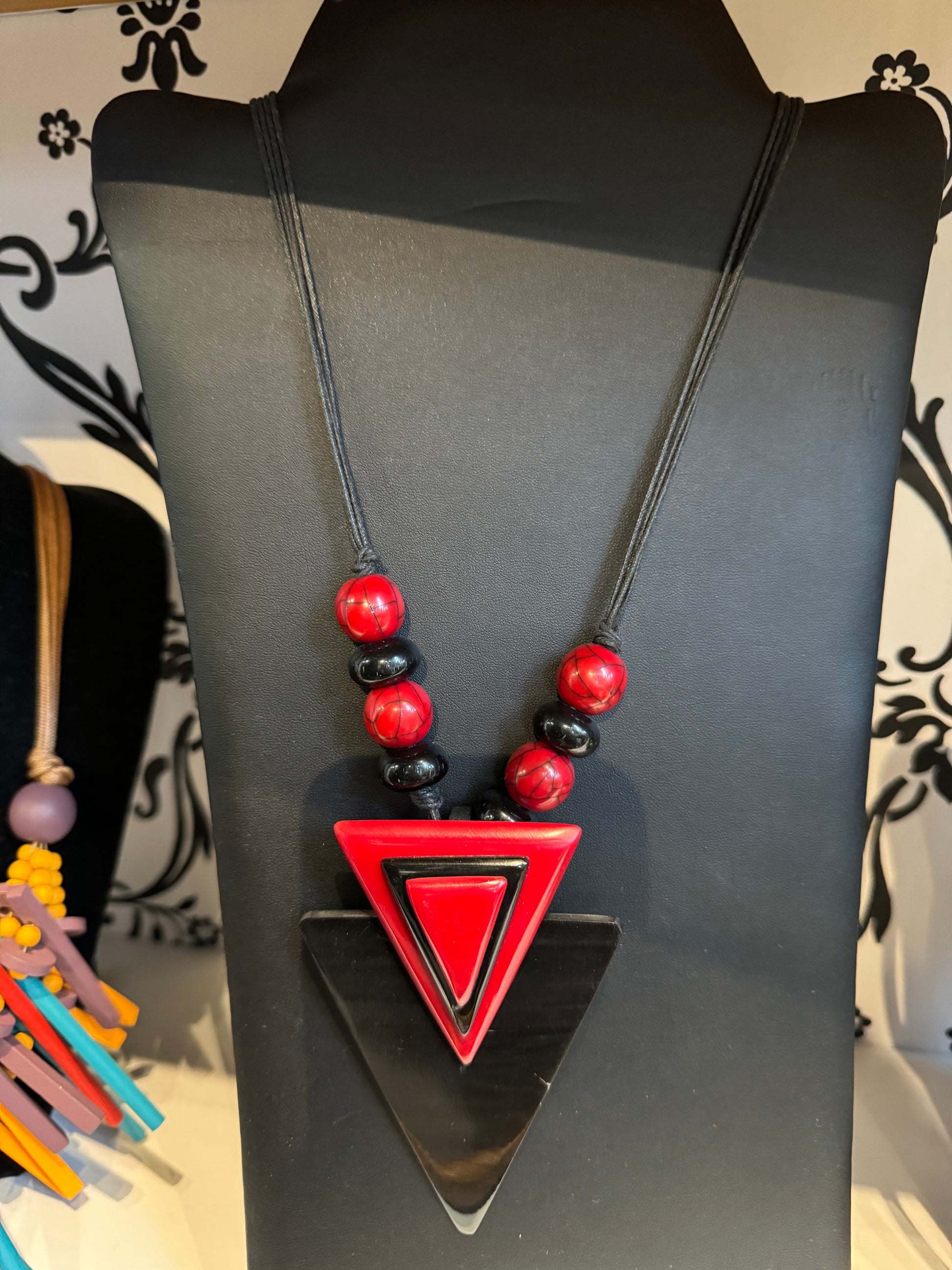 An elegant accessory displayed on a black stand, this bold look features a large triangular pendant with red and black layered triangles. Above the pendant are five round beads in alternating red and black colors. A Black and Red Necklace by Posies Flowers & Fashion Inc. is partially visible to the left.