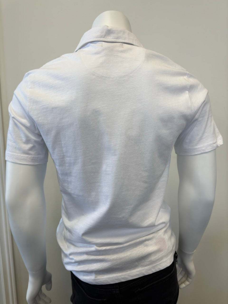 A mannequin wearing the Cotton Polo Men’s in white, featuring a classic collar and short sleeves, with three dark buttons on the placket, stands against a plain, light-colored background. The outfit is completed with dark pants from Silver Jeans Co., showcasing a casual yet stylish look.