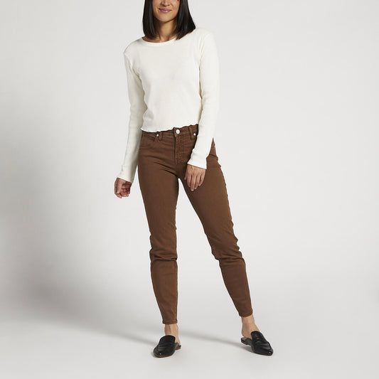 A person wearing a long-sleeved white shirt, Cecilia Mid Rise Skinny Pants - JAG in brown, and black slip-on shoes stands against a plain white background. They are slightly turned to the right and have one arm resting casually by their side.