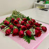 A bouquet of 2 Dozen Red Roses Freshcut by Posies Flowers & Fashion, accented with baby's breath and green leaves, rests on a pink mat atop a white table. Scissors and floral supplies can be seen in the background on another table.