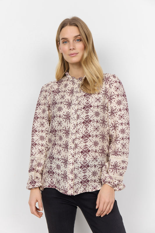 A woman with long blonde hair is wearing the luxurious blend of fabrics in the Shirt SC-IANE 2 from RAGWEAR, featuring a cream-colored blouse with a maroon floral pattern and black pants. She stands against a white background, looking directly at the camera with a neutral expression.