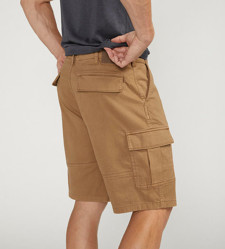 A man stands against a plain background wearing a dark gray t-shirt with a chest pocket, relaxed fit CARGO SHORT DESERT styled by Silver Jeans Co. (product code: M53151CBT639), and black sneakers with white soles. He appears to be adjusting his shirt with his right hand.